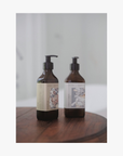 Lifestyle: Two amber glass pump bottles of La-Eva Shampoo and Conditioner with a label featuring 'Younger Together' artwork by Valeska Hykel standing on a dark wooden surface against a soft light gray background.