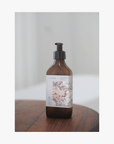 A recycled amber glass pump bottle of La-eva Roseum Hand & Body lotion with a label featuring 'Younger Together' artwork by Valeska Hykel and the text 'roséum ', part of the rose geranium botanical, Pelargonium roseum.