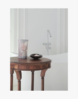 Lifestyle: A small dark ceramic dish containing La-Eva roseum bath salts sits on a round wooden table, with Valeska Hykel salt package visible beside it