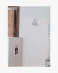 Lifestyle: Bathroom with shower head on wall and shower niche in corner with three pump bottles of Roseum Shampoo and Conditioner with a labels featuring 'Flamingos' artwork by Meinke Flessemen and the text 'roséum' part of the rose geranium botanical, Pelargonium roseum.