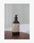 Lifestyle: an amber glass pump bottle of La-Eva Roseum Shampoo with a label featuring 'Younger Together' artwork by Valeska Hykel standing on a dark wooden surface against a soft light gray background.
