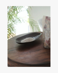 Lifestyle: A small dark ceramic dish containing La-Eva spicey bath salts sits on a round wooden table, with a floral-patterned container partially visible beside it and blurred green leaves in the background.