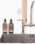 Lifestyle: Two recycled amber glass pump bottles of Roseum Hand & Body Wash and Lotion on kitchen sink side with bespoke artwork for ARC architecture by artist Beatrice Hassel McCosh.