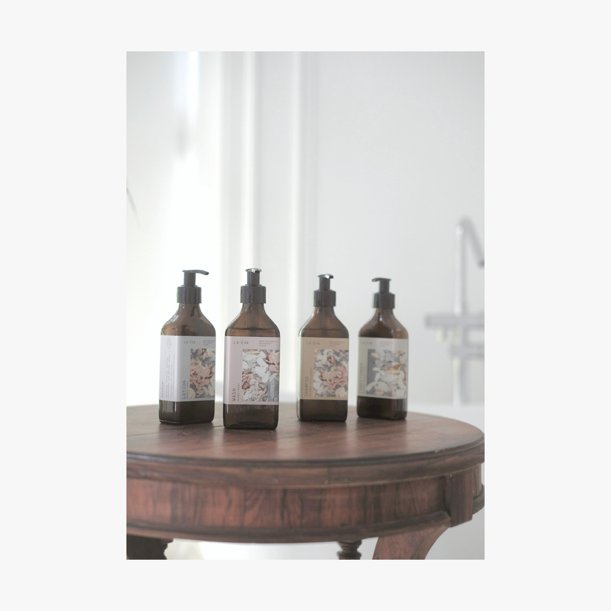Four amber glass pump La-Eva bottles with Valeska Hykel limited edition labels are lined up on a round wooden table against a softly blurred bathroom background.