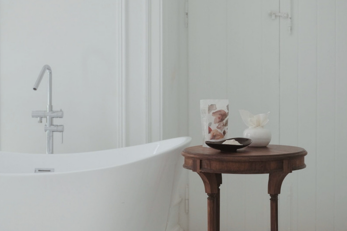 A white freestanding bathtub in a bright, minimalist bathroom with white panelled walls, a small wooden side table holding a La-Eva inhalation pomegranate and Valeska Hykel limited edition bath salts