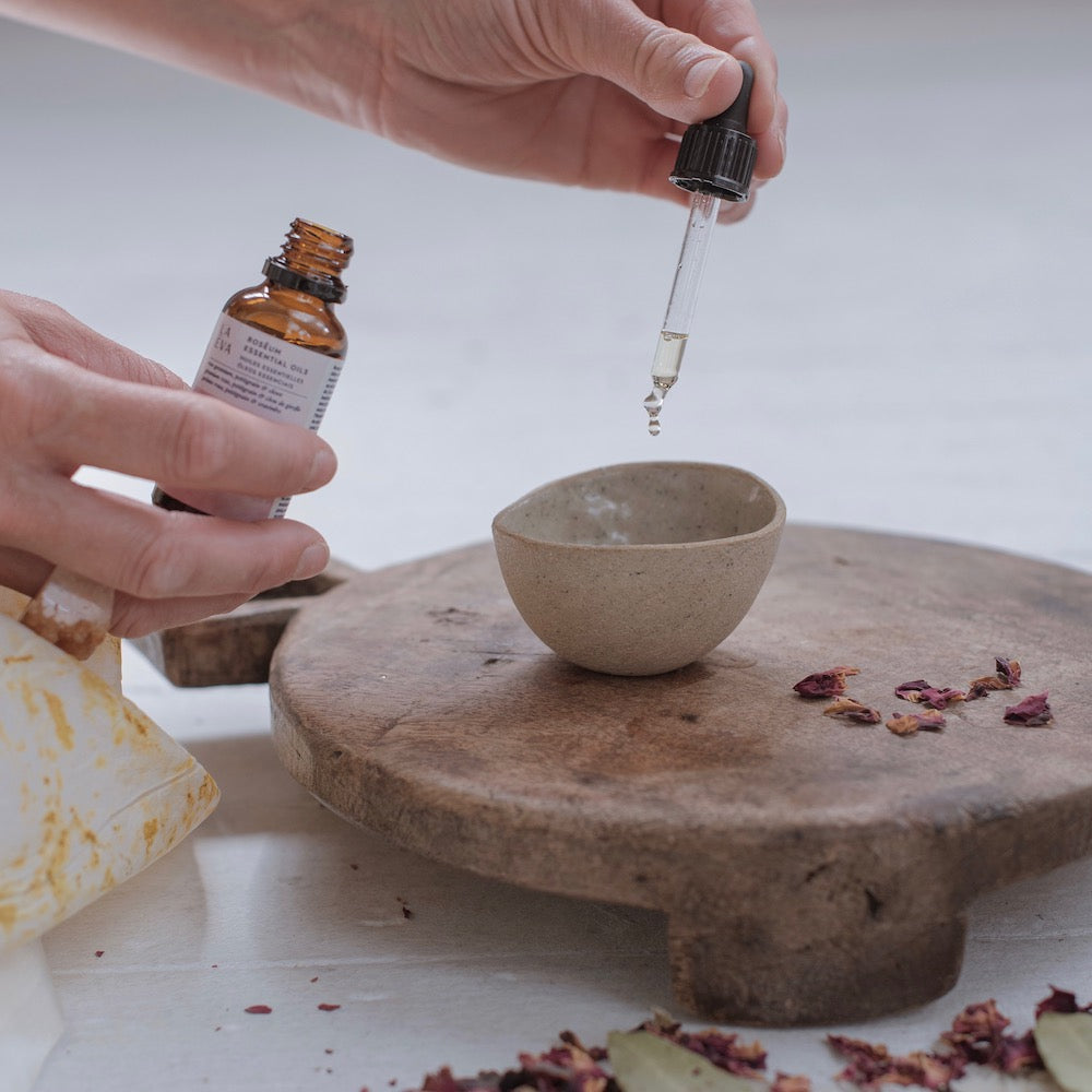 woman using a pipette to drop La-Eva Roseum essential oil into a small ceramic bowl resting on a wooden trestle, with dried rose petals scattered on the ground