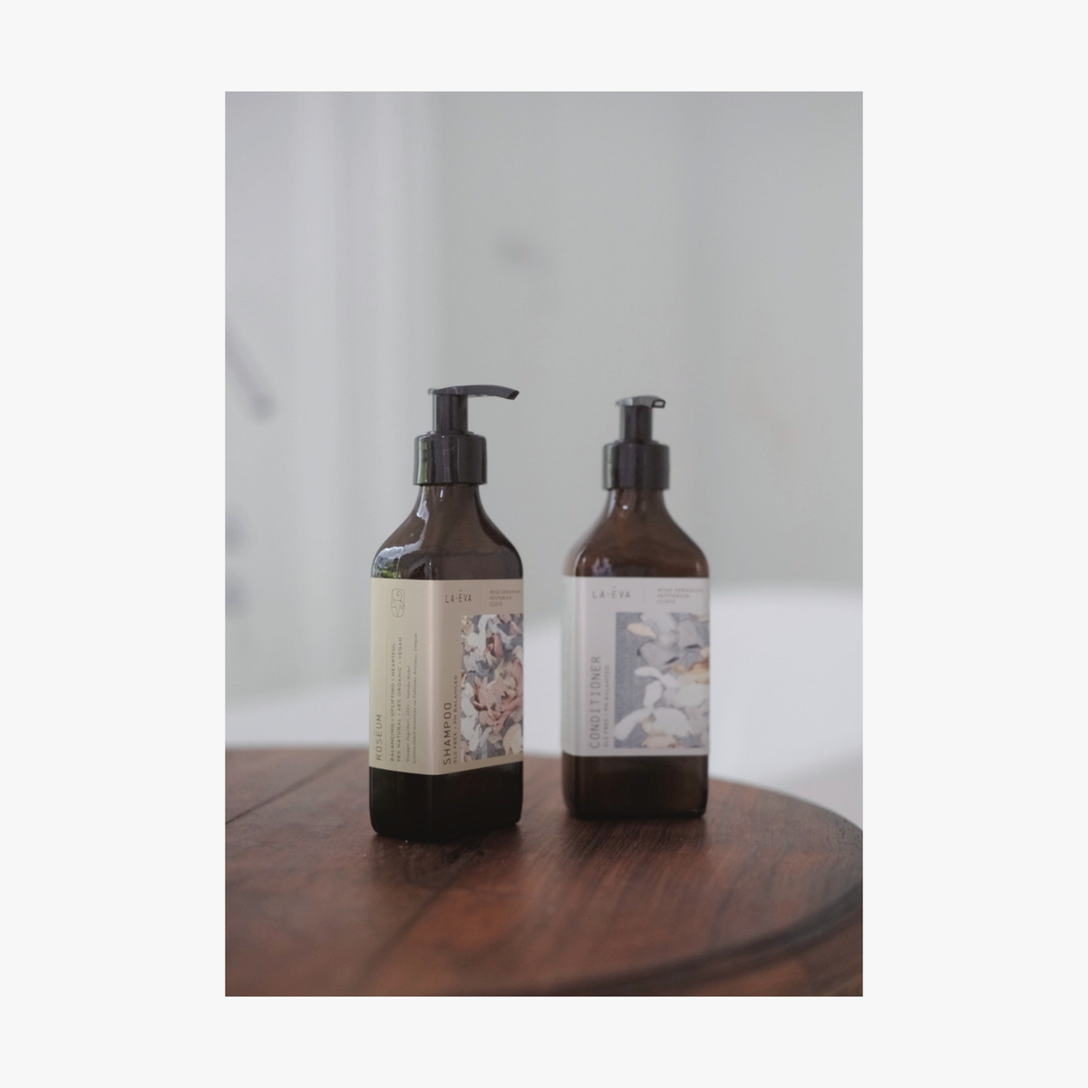 Lifestyle: Two amber glass pump bottles of La-Eva Shampoo and Conditioner with a label featuring 'Younger Together' artwork by Valeska Hykel standing on a dark wooden surface against a soft light gray background.