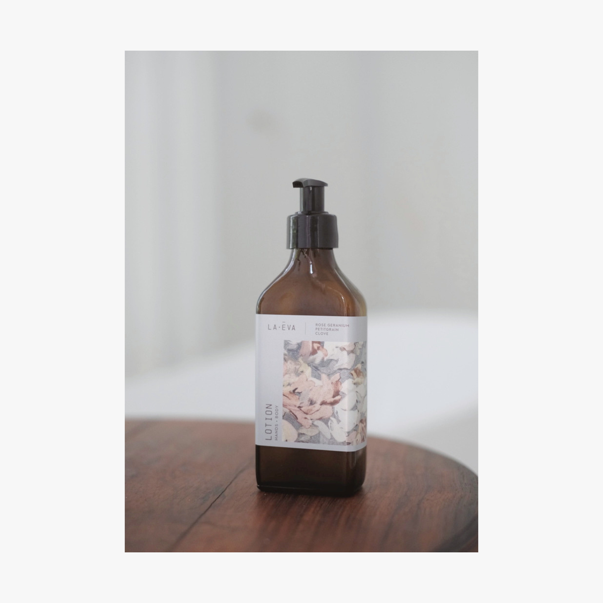 A recycled amber glass pump bottle of La-eva Roseum Hand & Body lotion with a label featuring 'Younger Together' artwork by Valeska Hykel and the text 'roséum ', part of the rose geranium botanical, Pelargonium roseum.