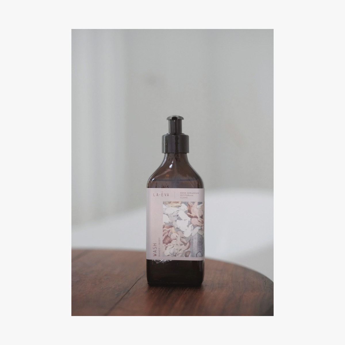 A recycled amber glass pump bottle of La-eva Roseum Hand & Body wash with a label featuring 'Younger Together' artwork by Valeska Hykel and the text 'roséum ', sitting on a wooden table with a soft background