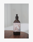 A recycled amber glass pump bottle of La-eva Roseum Hand & Body wash with a label featuring 'Younger Together' artwork by Valeska Hykel and the text 'roséum ', sitting on a wooden table with a soft background