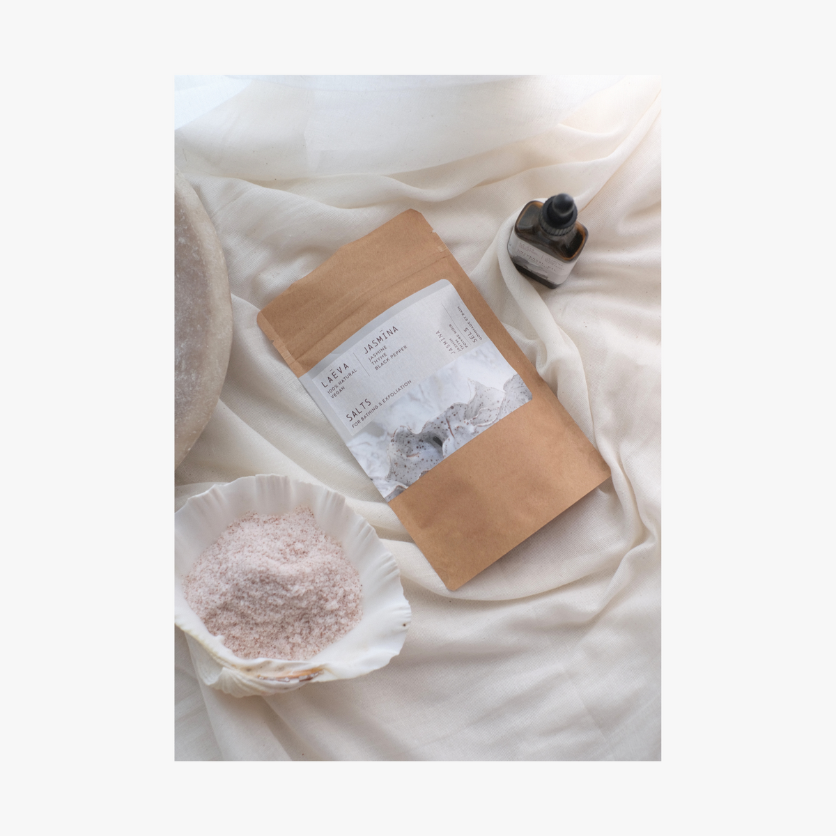 LIfestyle: A shell holding jasmina bath salts A package of La Eva Jasmina Exfoliant & Bath Salts with a label featuring artwork by Katie Coston, and jasmina universal oil on a bed