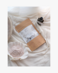 LIfestyle: A shell holding jasmina bath salts A package of La Eva Jasmina Exfoliant & Bath Salts with a label featuring artwork by Katie Coston, and jasmina universal oil on a bed