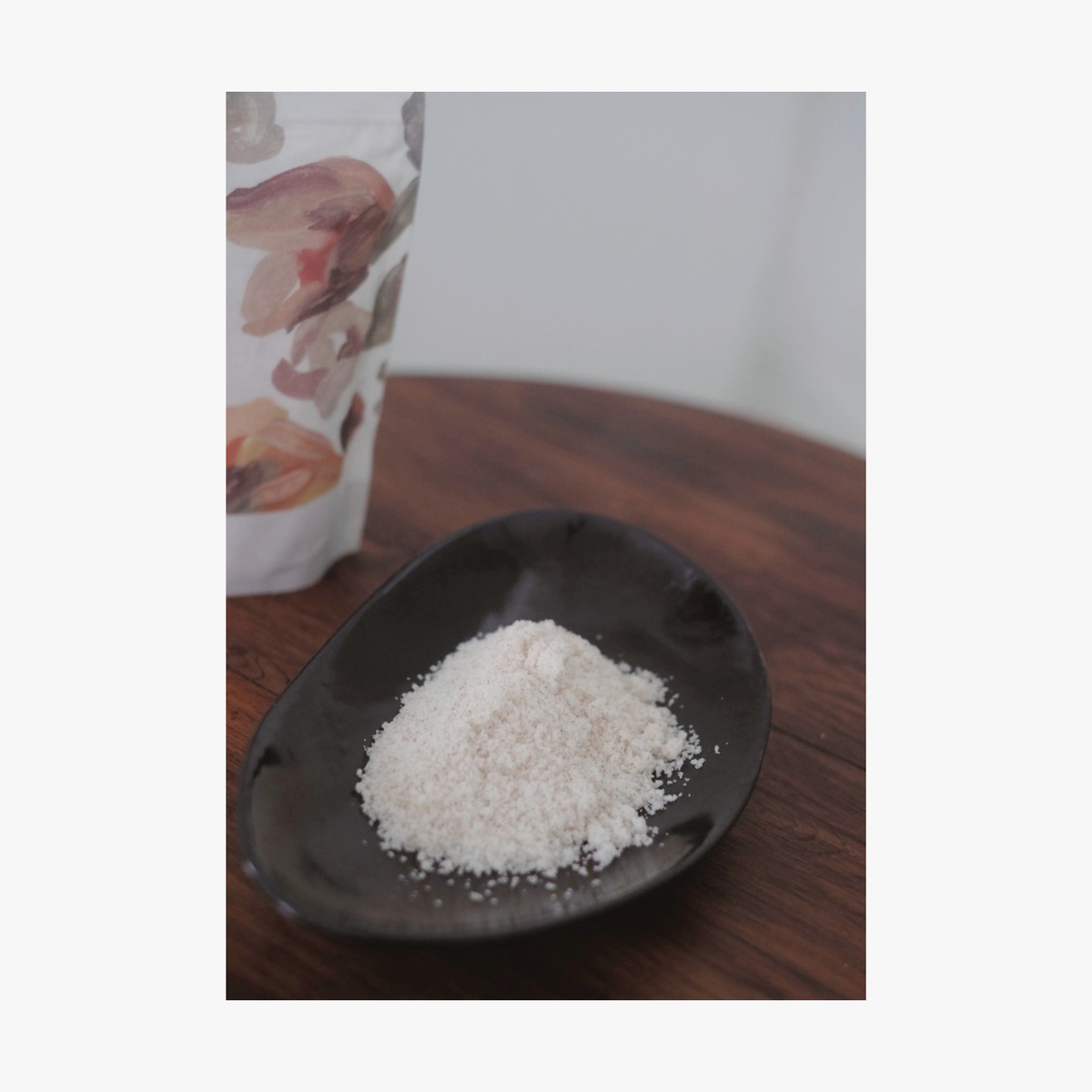 Lifestyle; A small dark ceramic dish containing La-Eva jasmina bath salts sits on a round wooden table, with Valeska Hykel salt package partially visible beside it