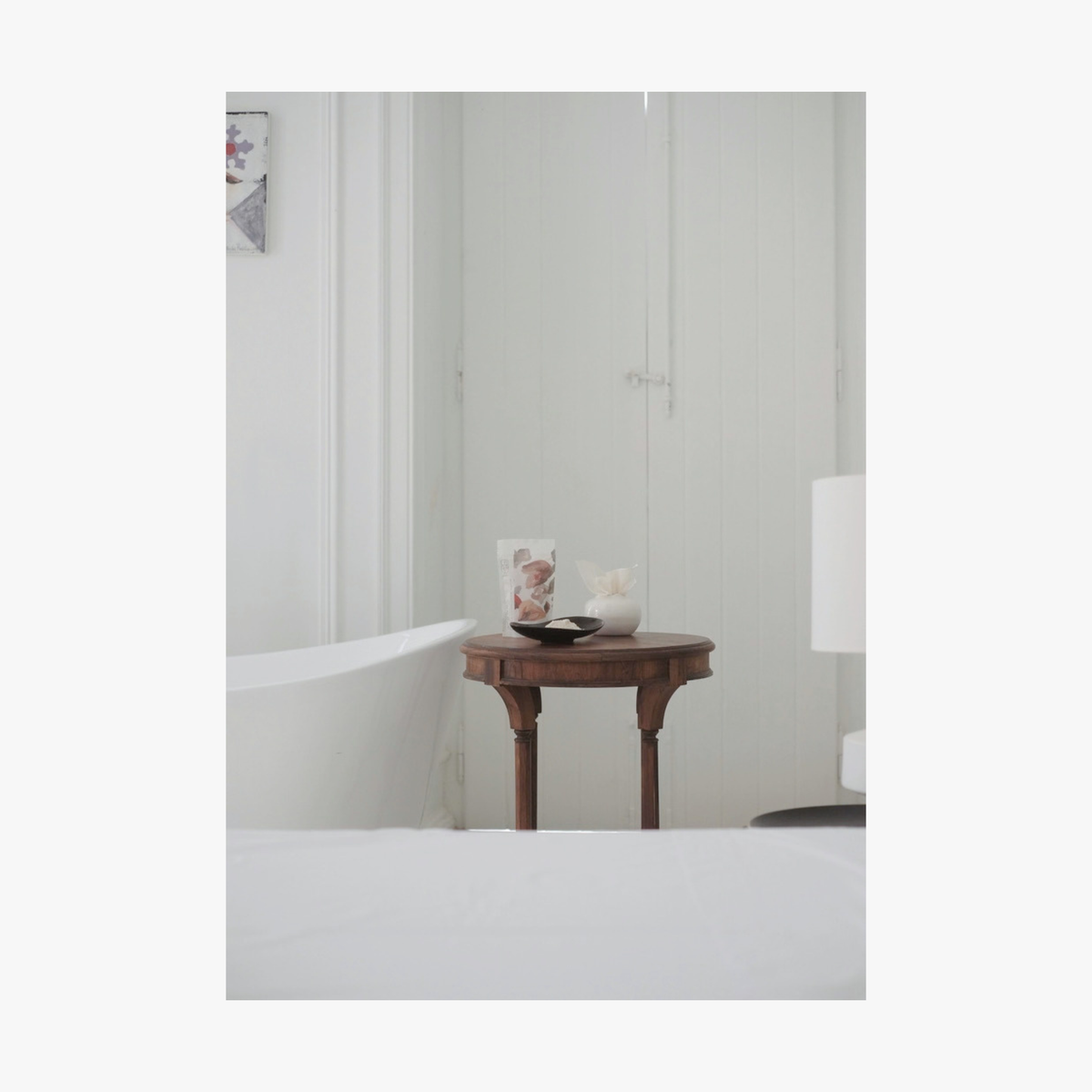 Lifestyle La-Eva: A styled interior scene with a small round wooden side table holding an inhilation pomegranate and a small dish of La-Eva jasmina salts, set beside a white bathtub and a white wall in a softly lit room.