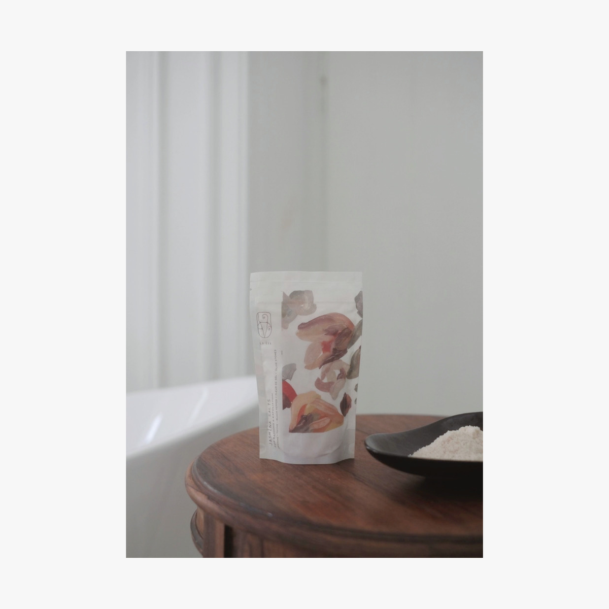 Lifestyle; A small dark ceramic dish containing La-Eva jasmina bath salts sits on a round wooden table, with Valeska Hykel salt package visible beside it