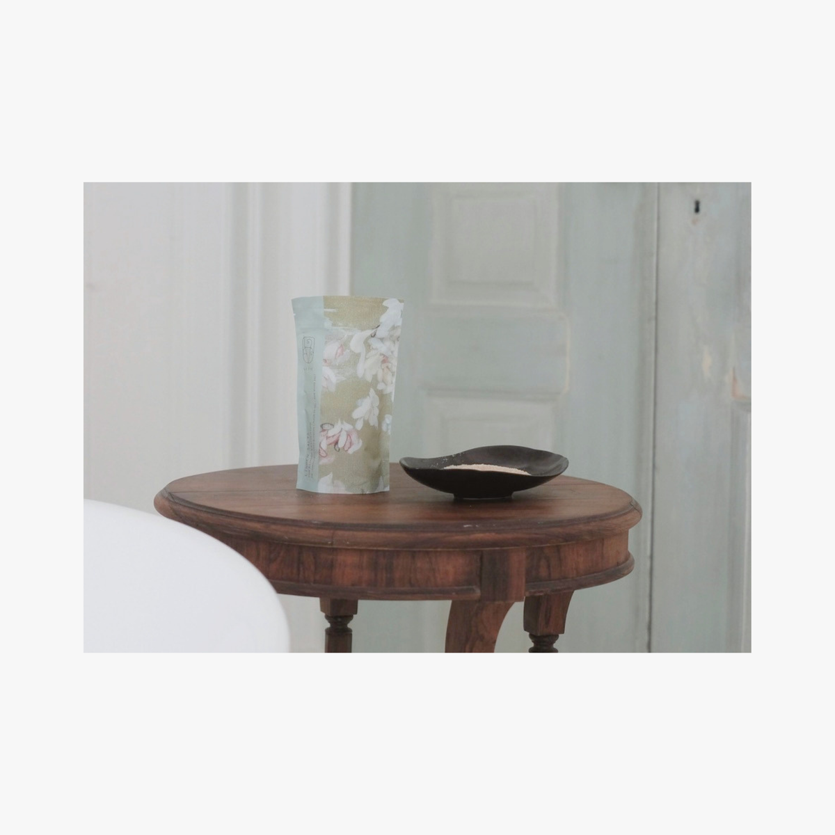 Lifestyle: A standing pouch of La-Eva Linden salts with Valeska Hykel artwork label sits on a round wooden table beside a small black dish in a softly lit interior setting