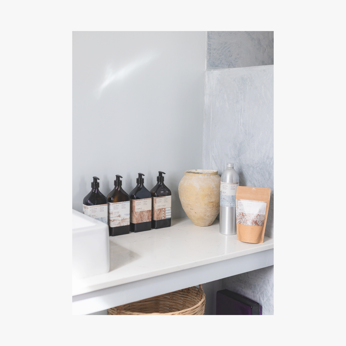 Bathroom shelf with bottles of the La-Eva refillable amenities spice collection and a vase against a light grey wall. Featuring artwork by Emma Cassi