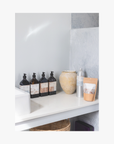 Bathroom shelf with bottles of the La-Eva refillable amenities spice collection and a vase against a light grey wall. 