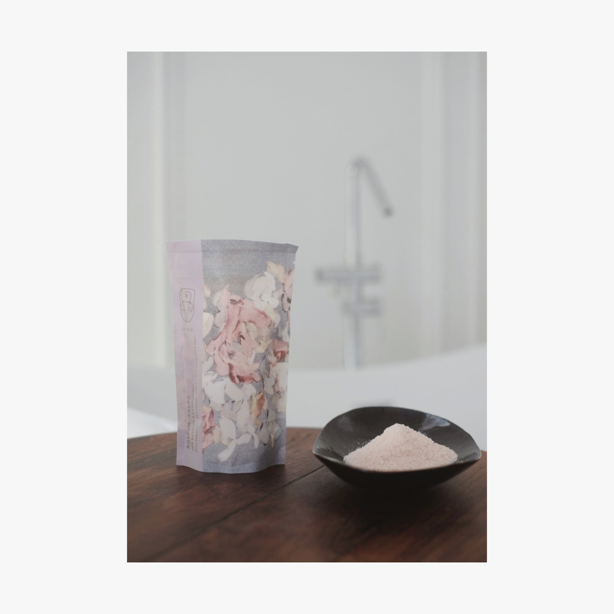 Lifestyle: A small dark ceramic dish containing La-Eva roseum bath salts sits on a round wooden table, with Valeska Hykel roseum salts package visible beside it