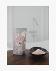 Lifestyle: A small dark ceramic dish containing La-Eva roseum bath salts sits on a round wooden table, with Valeska Hykel roseum salts package visible beside it