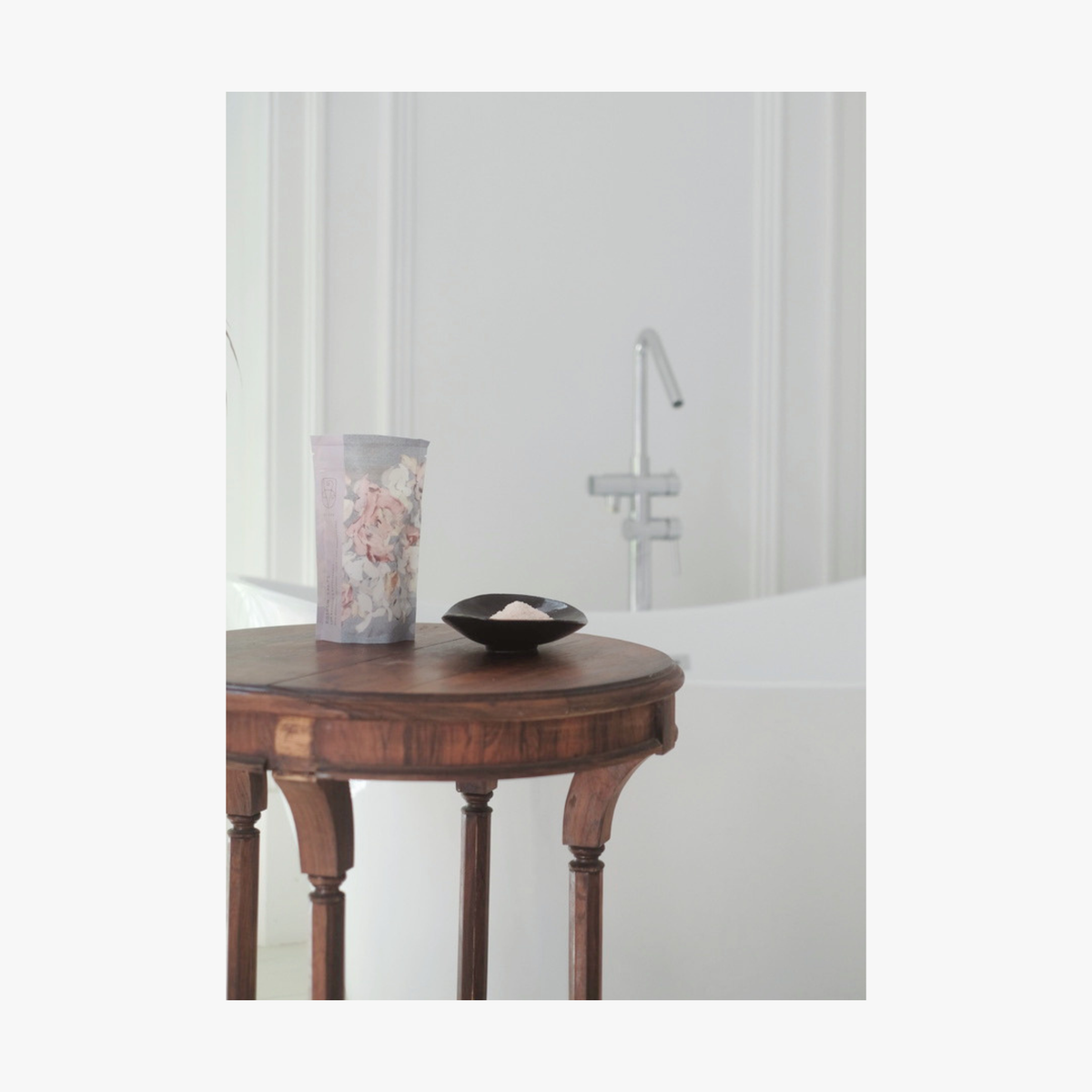 Lifestyle: A small dark ceramic dish containing La-Eva roseum bath salts sits on a round wooden table, with Valeska Hykel salt package visible beside it