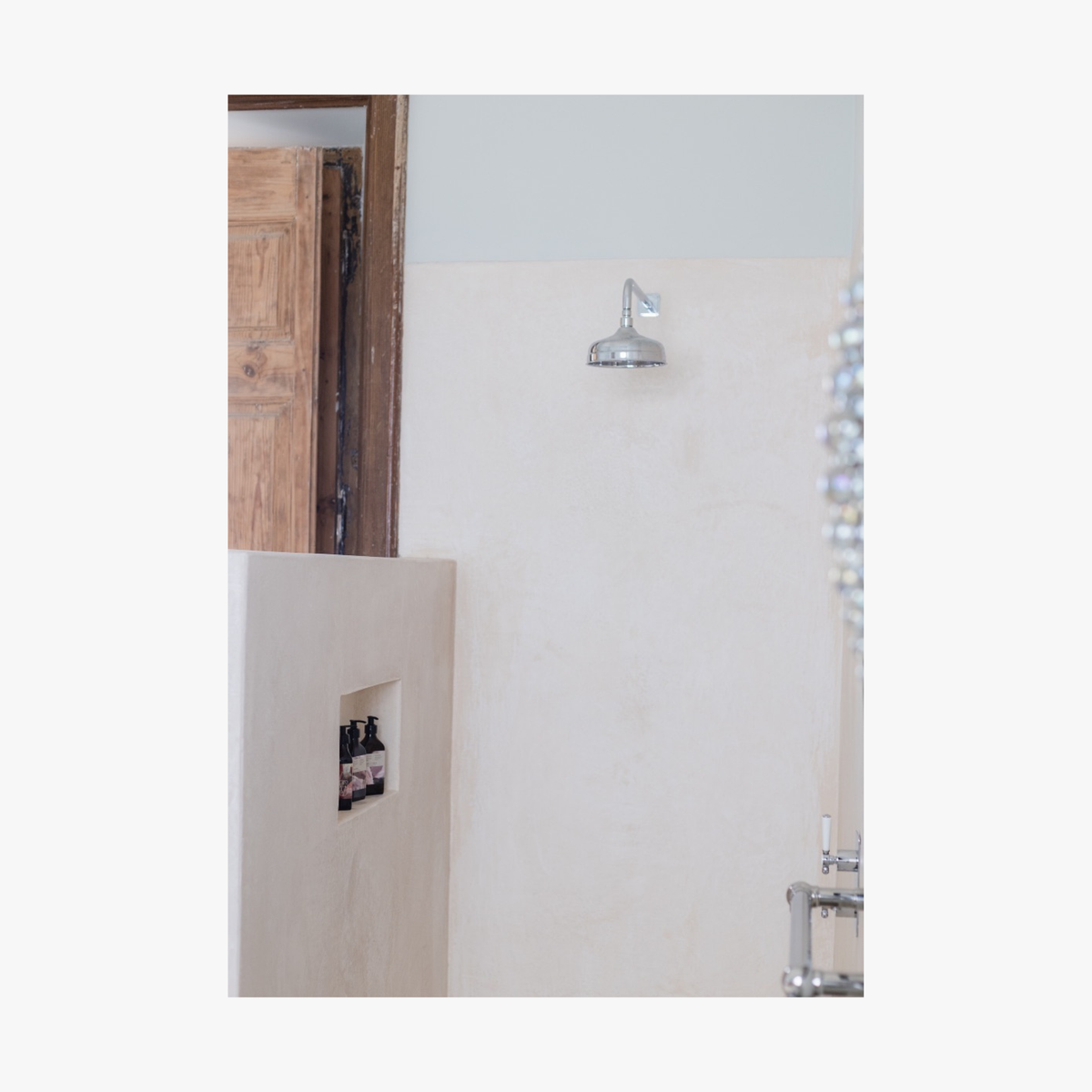 Lifestyle: Bathroom with shower head on wall and shower niche in corner with three pump bottles of Roseum Shampoo and Conditioner with a labels featuring 'Flamingos' artwork by Meinke Flessemen and the text 'roséum' part of the rose geranium botanical, Pelargonium roseum.