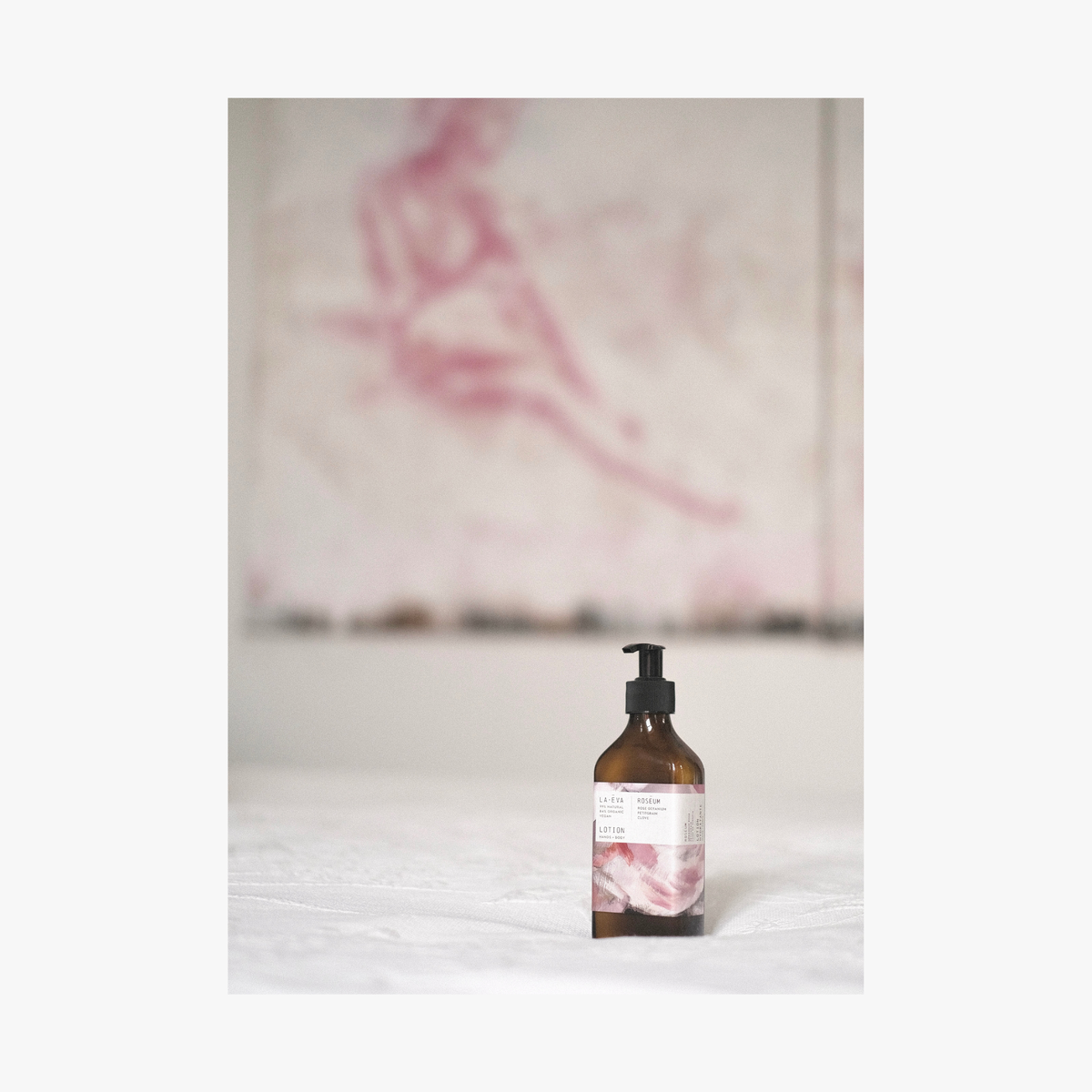 Lifestyle: a recycled amber glass bottle of roseum hand and body lotion with featuring labels with art by Meinke Flessemen on a white surface, with artwork in the background