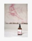 Lifestyle: a recycled amber glass bottle of roseum hand and body lotion with featuring labels with art by Meinke Flessemen on a white surface, with artwork in the background