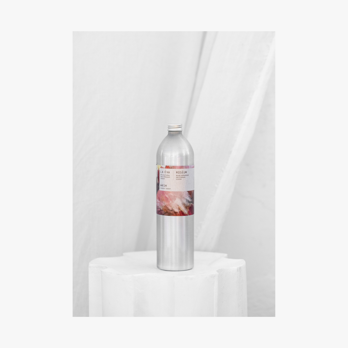 Lifestyle:A tall, slender aluminium bottle of Roseum hand and body wash sustainable refill. Label featuring Meinke Flessemen artwork.
