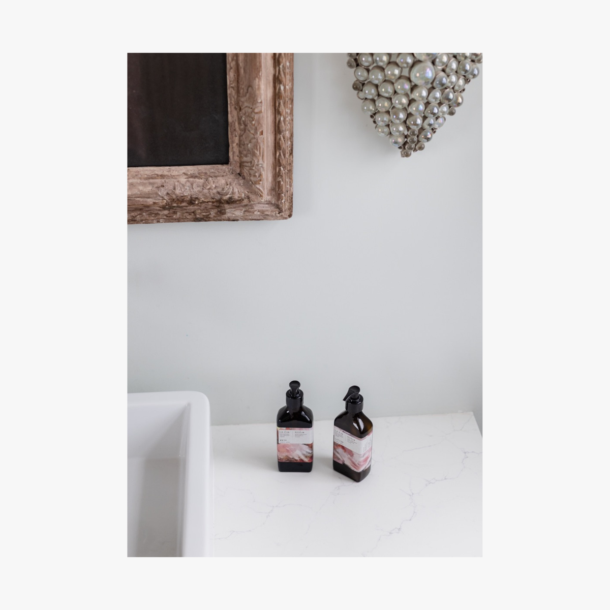 Two recycled amber glass bottles of roseum hand and body lotion with featuring labels with art by Meinke Flessemen on a white surface, framed mirror and decorative wall piece in the background.