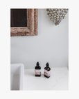 Two recycled amber glass bottles of roseum hand and body lotion with featuring labels with art by Meinke Flessemen on a white surface, framed mirror and decorative wall piece in the background.