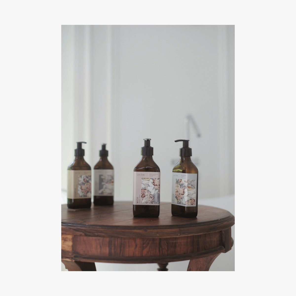 Three amber glass pump bottles of La-Eva Roseum collection with botanical Valeska Hykel labels are arranged on a round wooden table against a softly blurred light gray background.