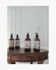 Three amber glass pump bottles of La-Eva Roseum collection with botanical Valeska Hykel labels are arranged on a round wooden table against a softly blurred light gray background.
