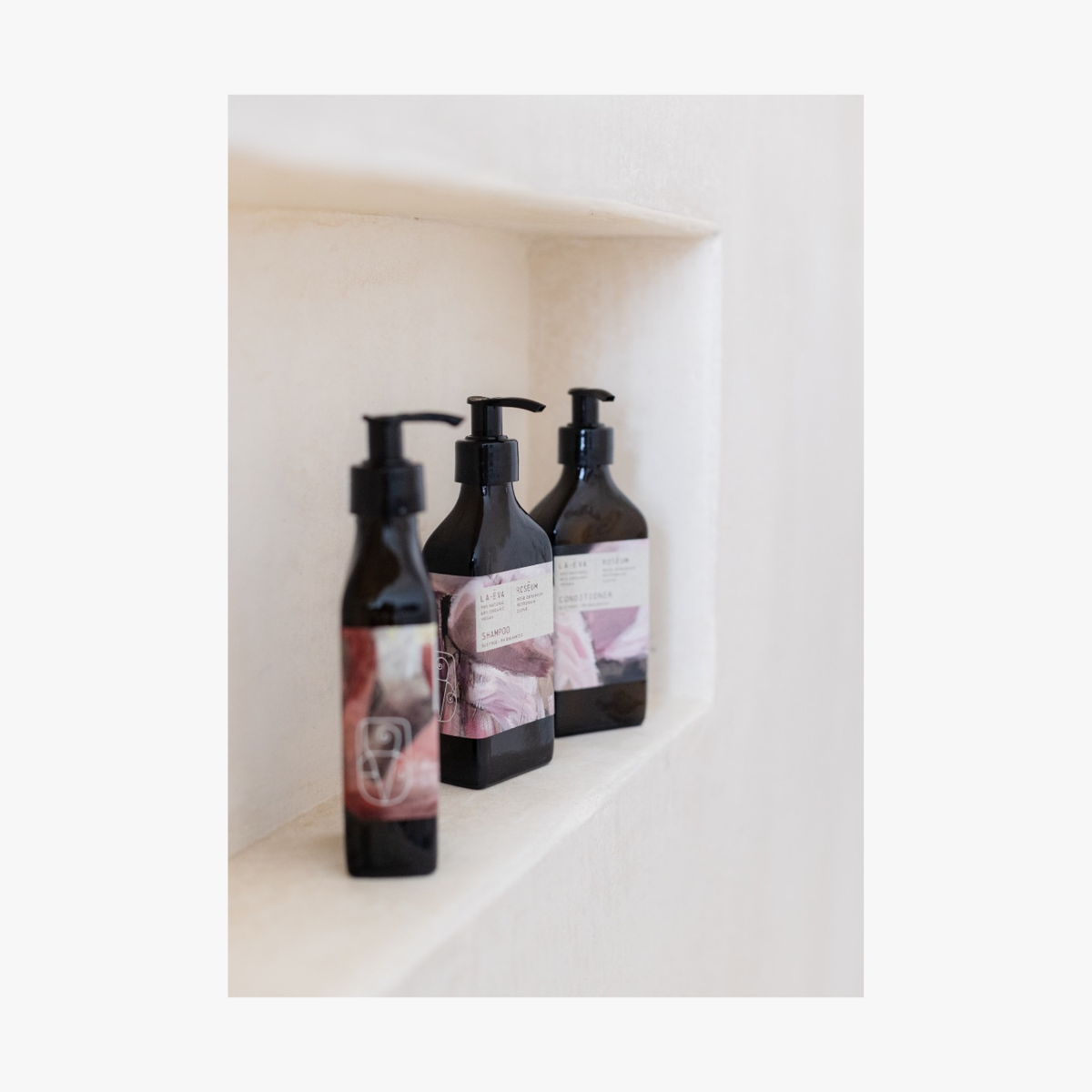 Lifestyle: three pump bottles of Roseum Shampoo and Conditioner with a labels featuring 'Flamingos' artwork by Meinke Flessemen and the text 'roséum' part of the rose geranium botanical, Pelargonium roseum.