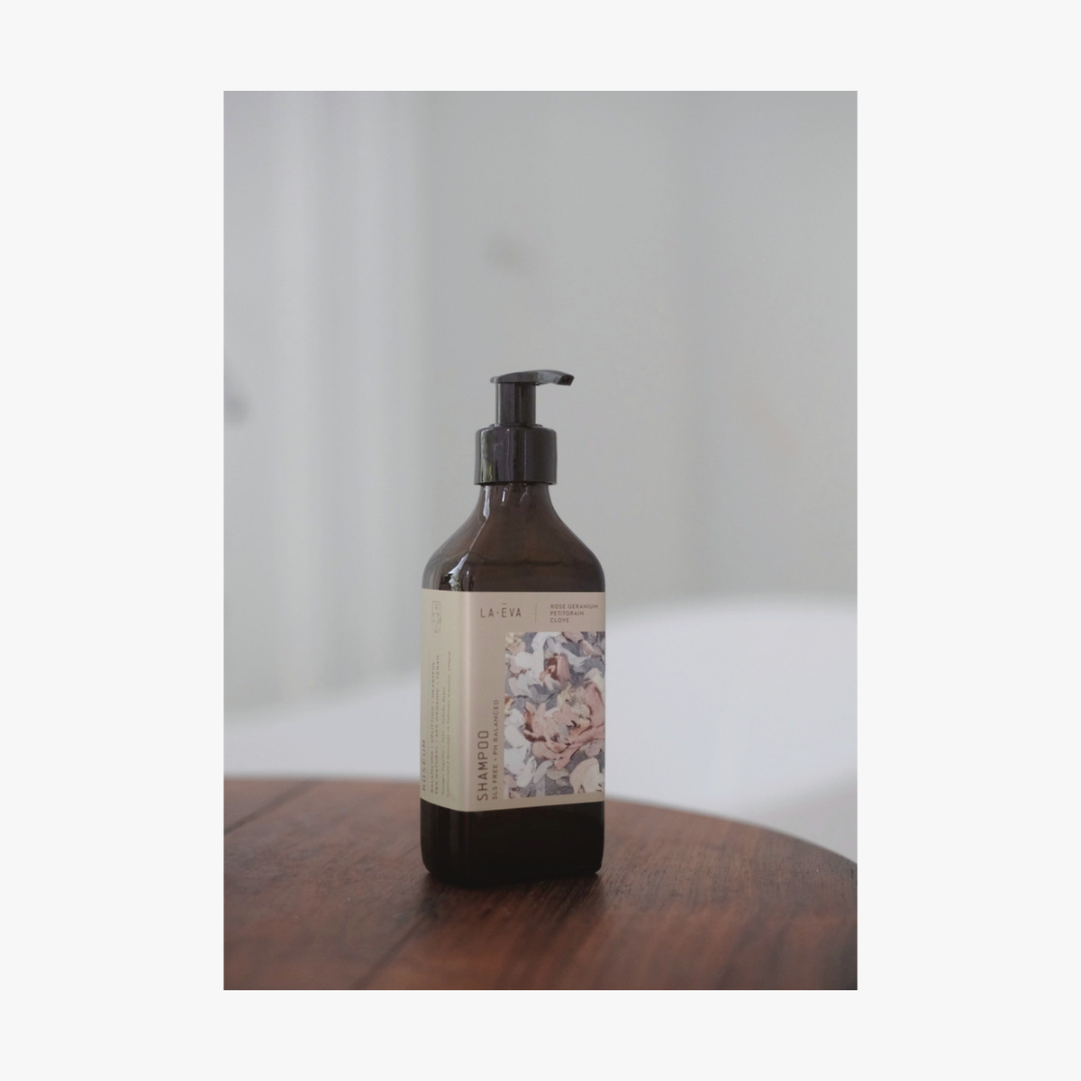 Lifestyle: an amber glass pump bottle of La-Eva Roseum Shampoo  with a label featuring 'Younger Together' artwork by Valeska Hykel standing on a dark wooden surface against a soft light gray background.