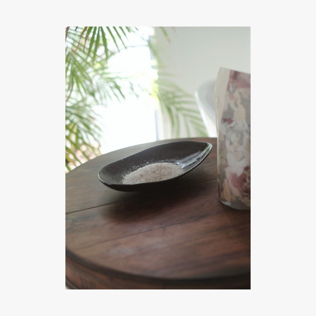 Lifestyle: A small dark ceramic dish containing La-Eva spicey bath salts sits on a round wooden table, with a floral-patterned container partially visible beside it and blurred green leaves in the background.