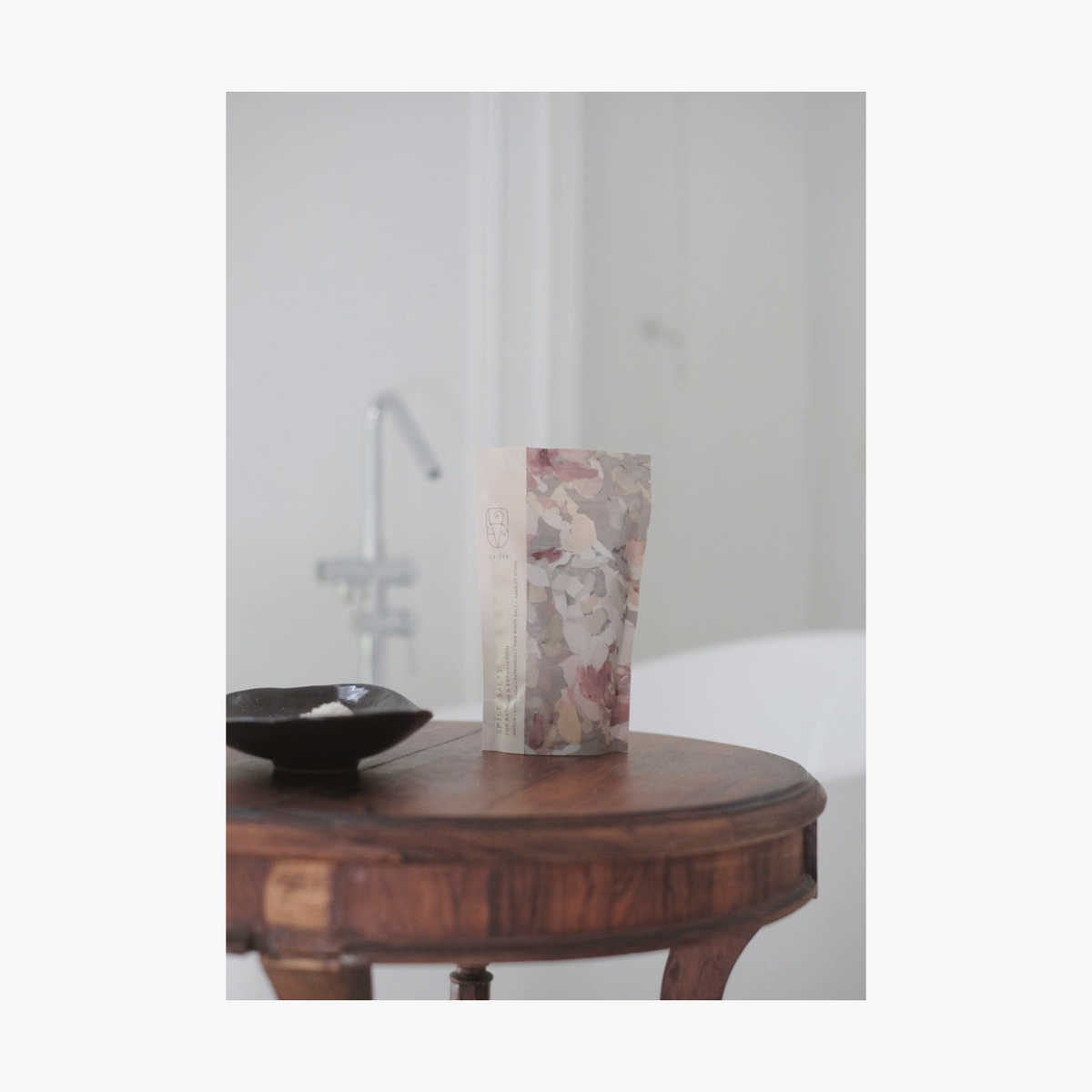 Lifestyle: A standing pouch of La-Eva spice salts with Valeska Hykel artwork label sits on a round wooden table beside a small black dish in a softly lit interior setting