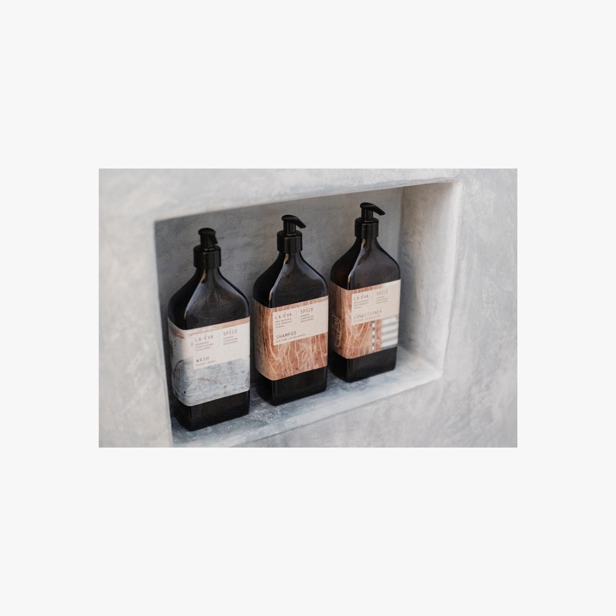 Three recycled amber glass pump bottles of La-Eva Spice shower collection with a labels featuring artwork by Emma Cassi,  Spicy wash; ginger, patchouli, cinnamon