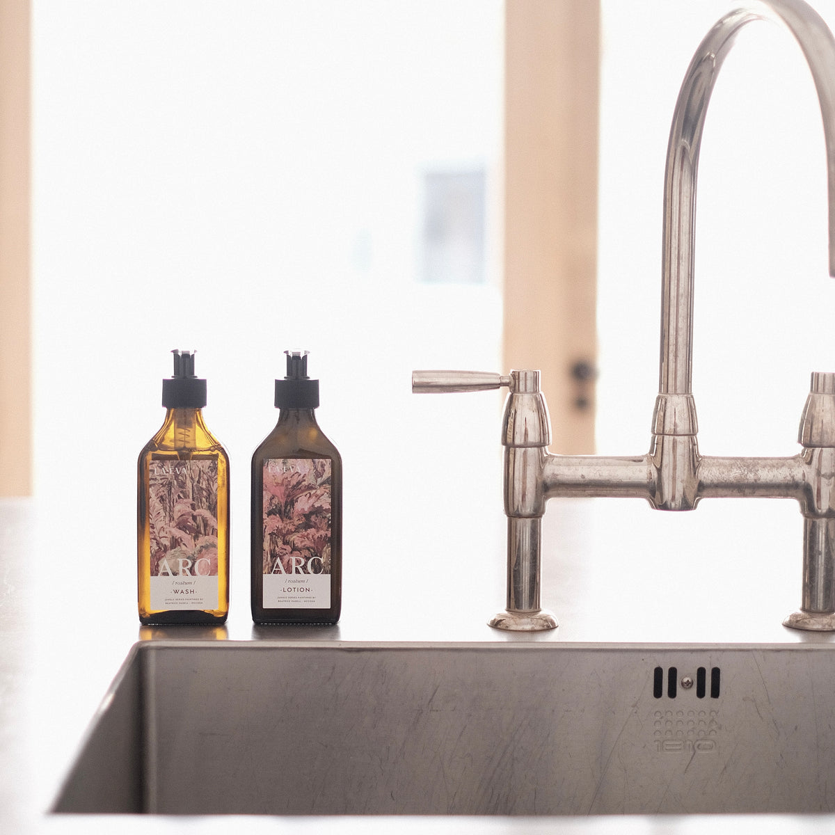 Lifestyle: Two recycled amber glass pump bottles of Roseum Hand & Body Wash and Lotion on kitchen sink side with bespoke artwork for ARC architecture by artist Beatrice Hassel McCosh. 