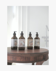 Four amber glass pump bottles with labels featuring artwork by Valeska Hykel are lined up on a round wooden table against a softly blurred bathroom background.