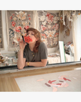 Valeska Hykel in her studio with original artwork in the background,leans over a table in a creative studio, holding a red flower to her mouth like a microphone. She is wearing a dark gray t-shirt and gold jewelry.
