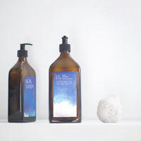 Two 500ml glass bottles of La-Eva Blu face and body lotion with a special edition label featuring the art of Nathalie Moisy, and a white ceramic pomegranate