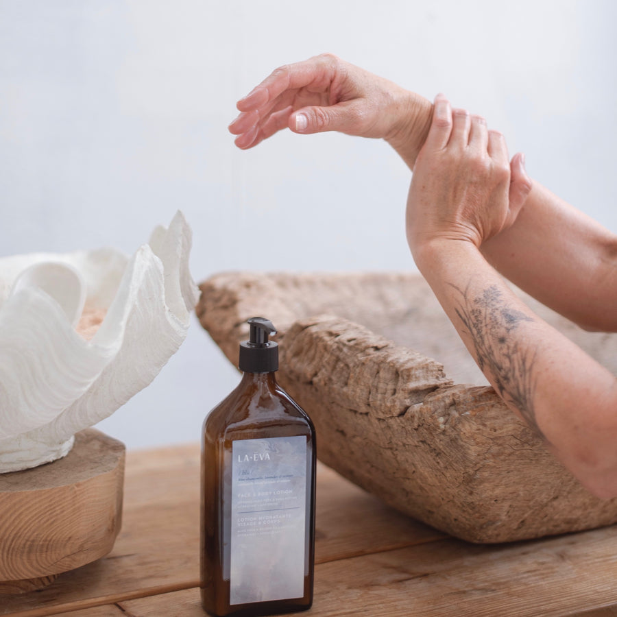 lady washing her hands beside a wooden sink, giant shell and a 500ml glass bottle of La-Eva Blu face and body wash
